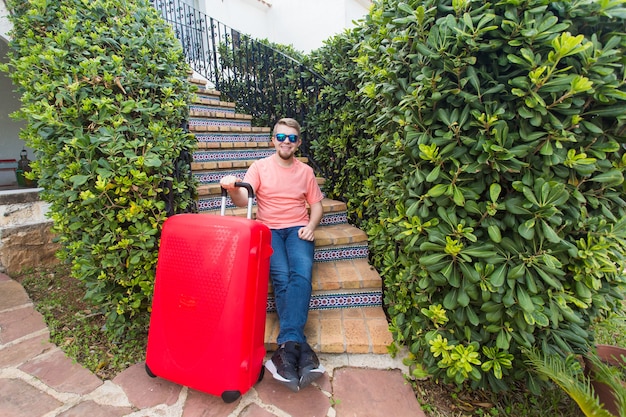 Concepto de vacaciones, viajes y personas. hombre con maleta de viaje roja sentado en las escaleras de su casa.