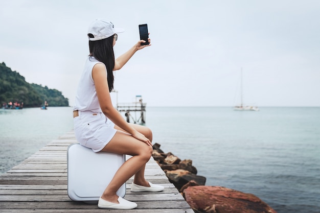 Concepto de vacaciones de viajes de verano, mujer asiática feliz viajero con teléfono móvil y maleta relajarse en el puente de madera en la playa del mar en Koh Kood, Trad, Tailandia