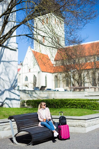 Concepto de vacaciones y viajes de verano - mujer joven sentada con café y maleta en el casco antiguo de Tallin, Estonia