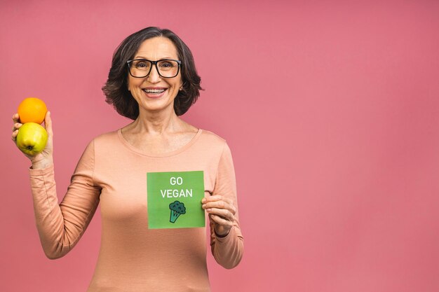 Concepto vegano o vegetariano. Retrato de una hermosa anciana madura mujer sosteniendo una manzana y naranja, sonriendo, aislado sobre fondo rosa. Una manzana al día mantiene alejado al médico.