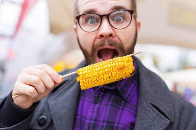 Concepto vegetariano y de comida - Hombre guapo comiendo maíz de comida callejera en el festival de comida rápida