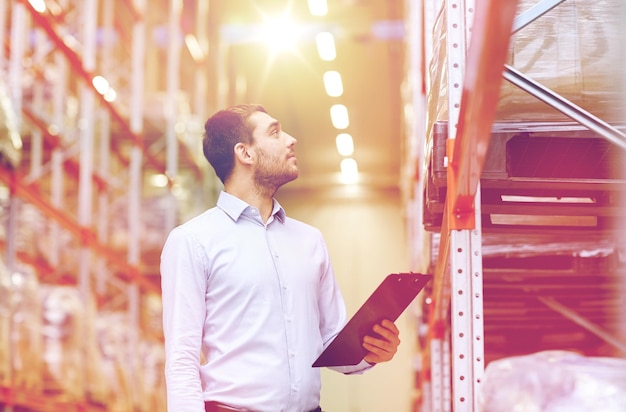 Foto concepto de venta al por mayor, logística, negocios, exportación y personas - hombre feliz o gerente con clipboard comprobando las mercancías en el almacén