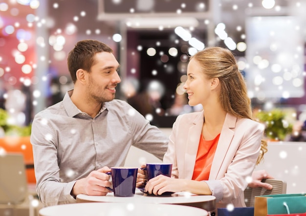 concepto de venta, compras, consumismo, ocio y personas - pareja feliz con bolsas de compras tomando café en el centro comercial con efecto de nieve