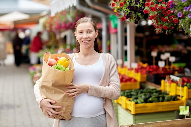 concepto de venta, compras, embarazo y personas - mujer embarazada feliz con una bolsa de papel llena de comida en el mercado callejero