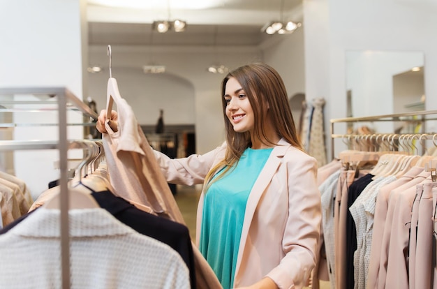 concepto de venta, compras, moda, estilo y personas - mujer joven feliz eligiendo ropa en el centro comercial o tienda de ropa