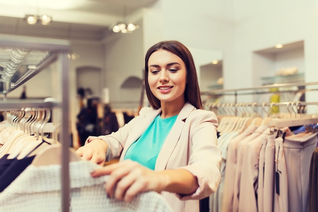 concepto de venta, compras, moda, estilo y personas: mujer joven feliz eligiendo ropa en el centro comercial o tienda de ropa