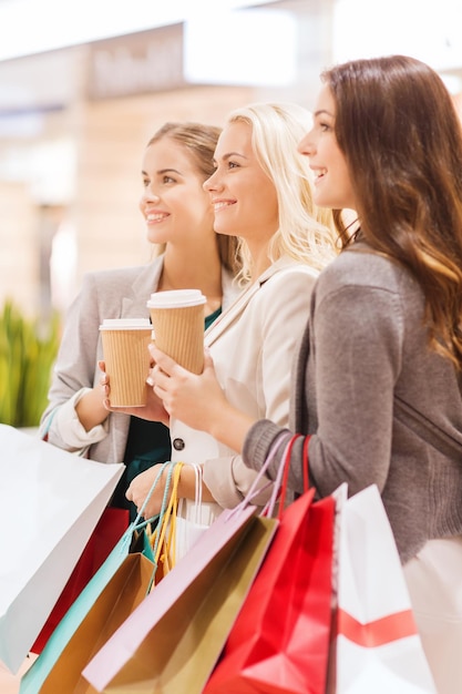 concepto de venta, consumismo y personas - mujeres jóvenes felices con bolsas de compras y taza de papel de café en el centro comercial