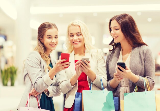 concepto de venta, consumismo, tecnología y personas - mujeres jóvenes felices con smartphones y bolsas de compras en el centro comercial