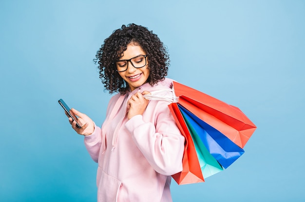 ¡Concepto de venta! Hermosa mujer afroamericana negra sonriendo y sosteniendo bolsas de la compra aisladas sobre fondo azul. Usando teléfono móvil.