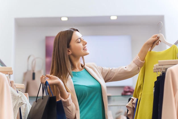 Concepto de venta, moda, consumismo y personas - mujer joven feliz con bolsas de compras eligiendo ropa en el centro comercial o tienda de ropa