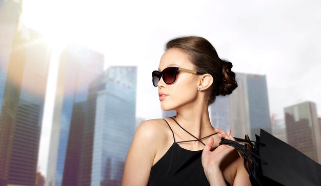 concepto de venta, moda, gente y lujo - mujer joven feliz y hermosa con gafas de sol negras con bolsas de compras sobre el fondo de los rascacielos de la ciudad