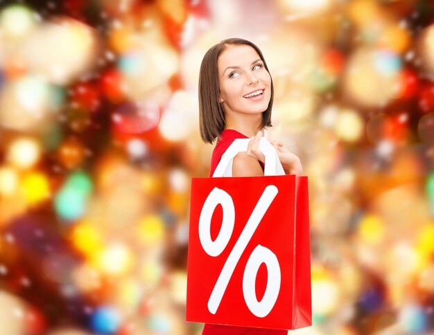 Foto concepto de venta, regalos, navidad, vacaciones y personas - mujer sonriente vestida de rojo con bolsas de compras y signo de porcentaje sobre fondo de luces rojas