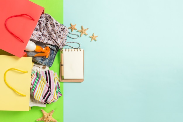 Concepto de verano de coloridas bolsas llenas de ropa. Bolsas de regalo con camiseta, shorts de mezclilla, cuaderno y botella de protector solar. Concepto de lista de deseos de verano. Espacio de copia de vista superior sobre fondo azul.