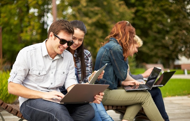 concepto de verano, educación, tecnología y personas - grupo de estudiantes o adolescentes con computadoras portátiles sentados en un banco al aire libre