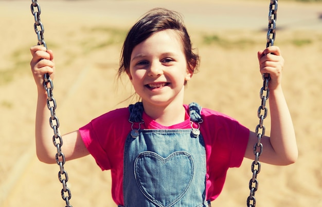 concepto de verano, infancia, ocio, amistad y personas - niñita feliz columpiándose en el patio de juegos para niños