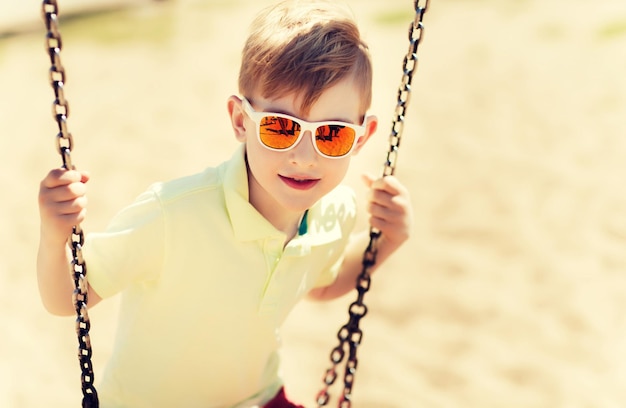 concepto de verano, infancia, ocio, amistad y personas - niño feliz con gafas de sol columpiándose en el patio de recreo de los niños