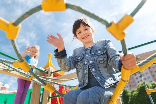 concepto de verano, infancia, ocio, gesto y personas - niñita feliz saludando con la mano en el marco de escalada del parque infantil