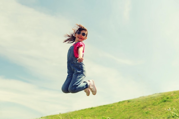 concepto de verano, infancia, ocio y personas - niña feliz saltando alto sobre el campo verde y el cielo azul al aire libre