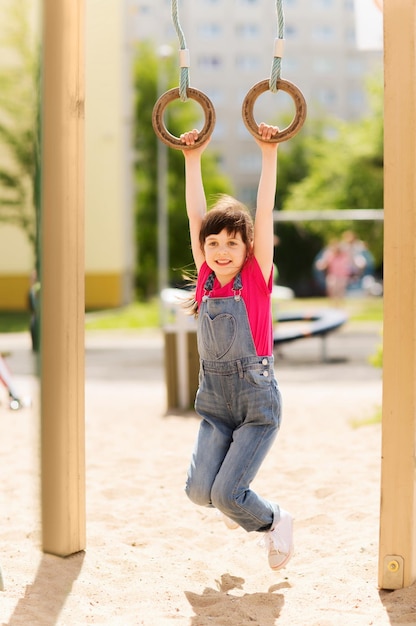 concepto de verano, infancia, ocio y personas - niñita feliz colgando de anillos de gimnasia en el parque infantil