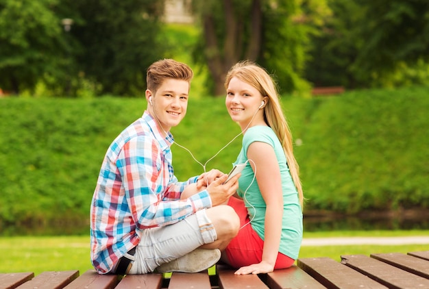 concepto de verano, vacaciones, vacaciones, tecnología y amistad - pareja sonriente con smartphone y auriculares sentados en un banco en el parque