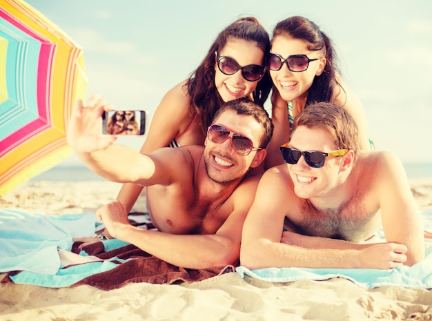 concepto de verano, vacaciones, vacaciones, tecnología y felicidad - grupo de personas sonrientes con gafas de sol tomando fotos con un smartphone en la playa