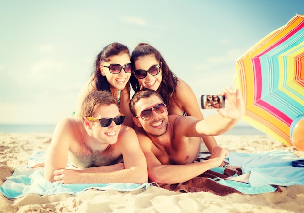 concepto de verano, vacaciones, vacaciones, tecnología y felicidad - grupo de personas sonrientes con gafas de sol tomando fotos con un smartphone en la playa