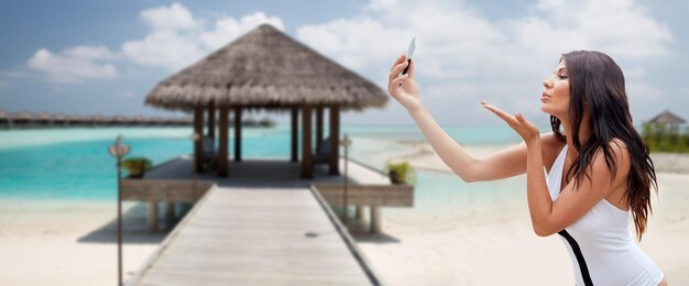 concepto de verano, viajes, tecnología y personas - mujer joven sexy tomando selfie con teléfono inteligente y enviando beso de golpe sobre bungalow en el fondo de la playa