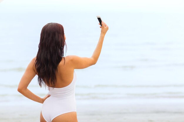 concepto de verano, viajes, tecnología y personas - mujer joven sexy tomando selfie con teléfono inteligente en la playa