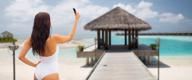 concepto de verano, viajes, tecnología y personas - mujer joven sexy tomando selfie con teléfono inteligente sobre bungalow en el fondo de la playa