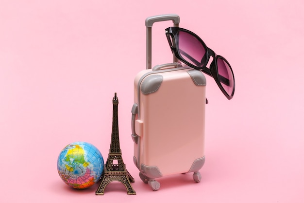 Concepto viajado. Mini maleta de viaje de plástico con gafas de sol y estatuilla de la Torre Eiffel, globo sobre fondo rosa.
