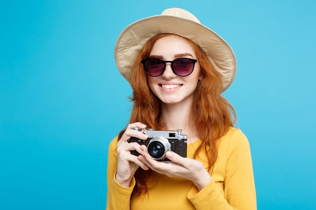 Concepto de viaje - Close up Retrato joven y bella chica atractiva redhair con sombrero de moda, gafas de sol y cámara vintage sonriendo a la cámara. Pared azul pastel. Copia espacio