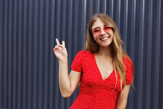 Concepto de viaje Hermosa chica hipster disfrutando de las vacaciones en las islas tropicales posando cerca de la muralla de la ciudad urbana con el signo de la paz sonriendo feliz a la cámara