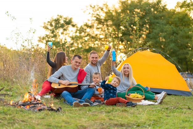 Concepto de viaje, turismo, caminata, picnic y personas - grupo de amigos felices con carpa y bebidas tocando la guitarra en el campamento