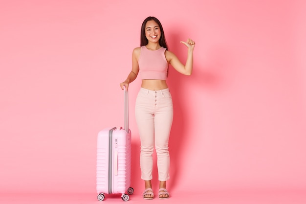 Concepto de viaje, vacaciones y vacaciones. Longitud total de una turista asiática descarada y feliz de pie sobre una pared rosa con una maleta y apuntando a sí misma, explorar países