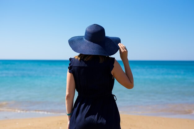 Concepto de viaje y vacaciones de verano - vista posterior de la mujer en vestido y sombrero sobre fondo de playa de verano