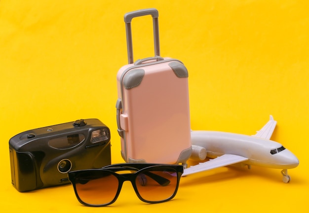 Concepto de viajes aéreos. Mini maleta de viaje de plástico rosa, avión, cámara y gafas de sol sobre fondo amarillo.