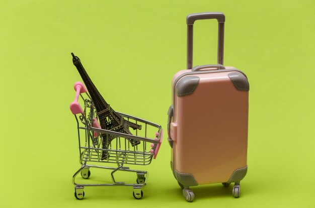 Concepto de viajes y compras. Mini maleta de viaje de plástico y carro de supermercado con estatuilla de la Torre Eiffel sobre fondo verde
