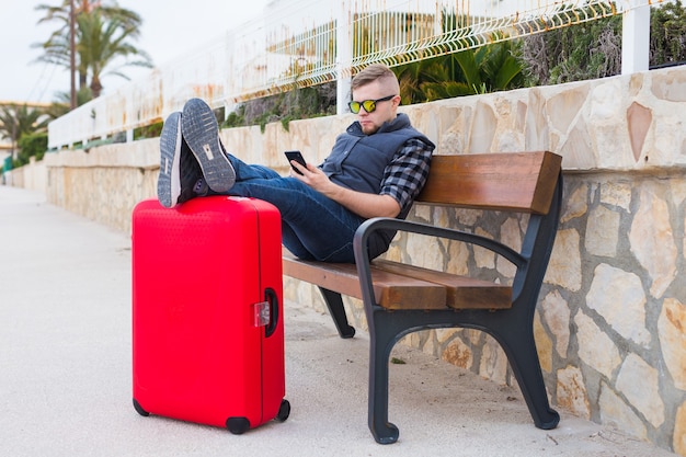 Concepto de viajes, turismo y personas. guapo se sienta y pone los pies en la maleta, mirando algo en su móvil.