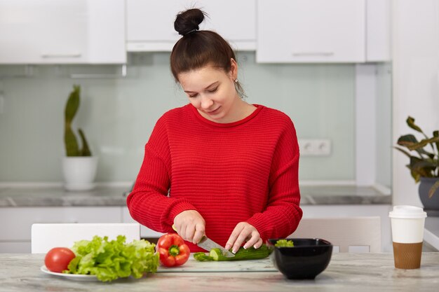 Concepto de vida doméstica y maternidad. Ocupada mujer embarazada corta verduras para hacer ensaladas, posa en el moderno apartamento contra el interior de la cocina, usa un suéter rojo, come sano