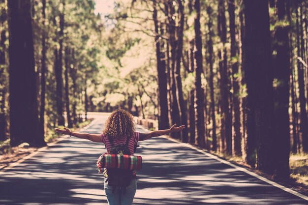 Concepto de vida de satisfacción de eternidad y éxito con chica solitaria rizado hai rin en medio de un largo camino en el bosque con los brazos abiertos y la mochila: pasión por los viajes y personas de estilo de viaje