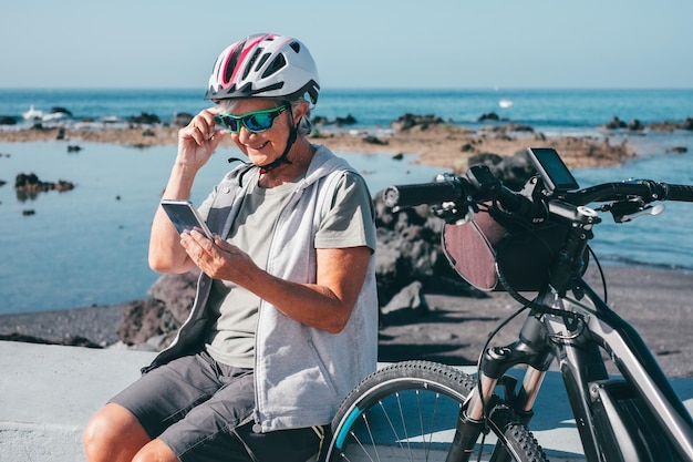 Concepto de videollamada Mujer ciclista senior sonriente sentada en la playa con bicicleta eléctrica usando el teléfono