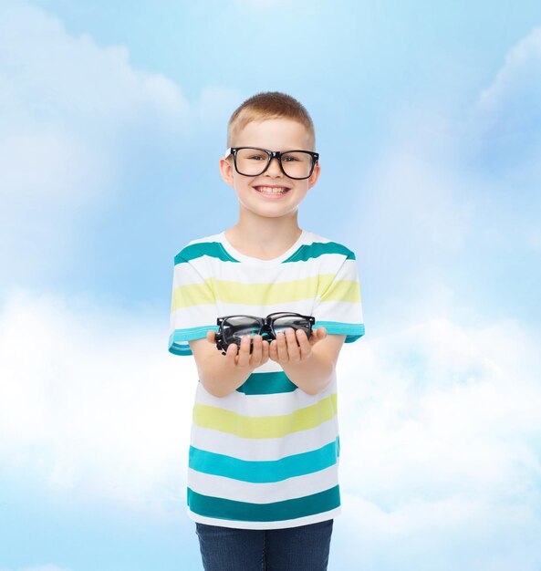 concepto de visión, salud y personas - niño sonriente con anteojos sosteniendo espectáculos sobre el cielo azul con fondo de nubes blancas