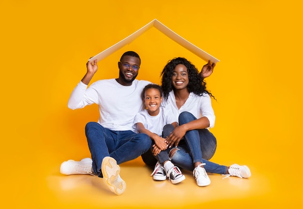 Concepto de vivienda familiar. Felices padres afroamericanos y su hija sentados bajo un techo simbólico soñando con un nuevo hogar, fondo amarillo