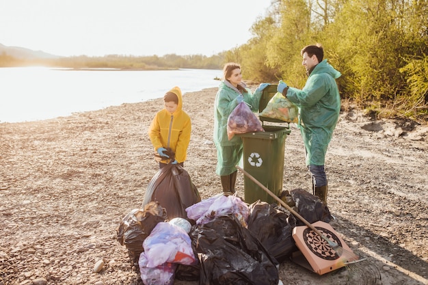 Concepto de voluntariado, caridad, limpieza, personas y ecología. Grupo de voluntarios familiares felices con bolsas de basura limpiando el área en el parque cerca del lago.