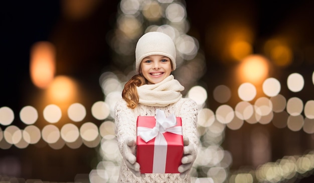 conceptos de vacaciones y personas - chica feliz en ropa de invierno con caja de regalos sobre luces de árbol de Navidad de fondo