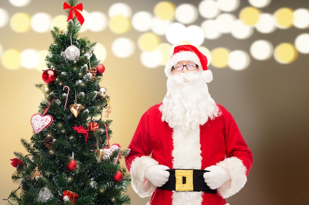 conceptos de vacaciones y personas - hombre disfrazado de Santa Claus en el árbol de Navidad sobre las luces de fondo