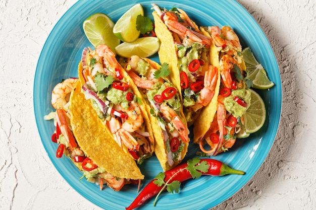 conchas de taco com camarões grelhados, salada de repolho, limão e guacamole em um prato azul sobre uma mesa branca, postura plana, espaço livre, close-up