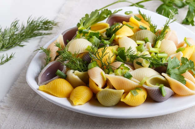 Conchiglie pasta coloreada con verduras frescas en un mantel de lino sobre madera blanca