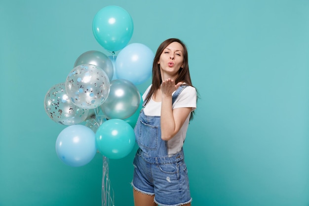 Concurso jovem em roupas jeans soprando enviando beijo de ar, comemorando e segurando balões de ar coloridos isolados no fundo da parede azul turquesa. festa de aniversário, conceito de emoções de pessoas.