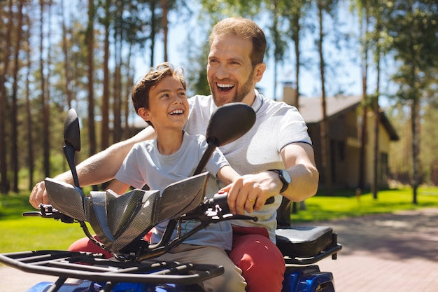 Conducción de velocidad. Padre joven inspirado sonriendo y conduciendo un vehículo todo terreno con su hijo